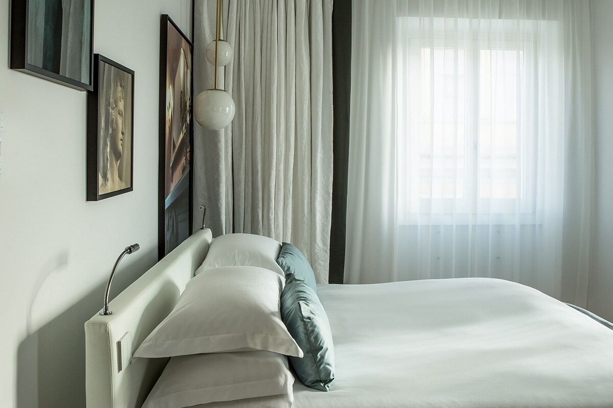 A hotel room with a large bed, a bedside table, a round bedside table, and a window with sheer curtains.