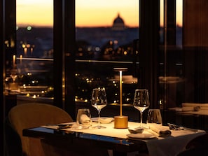 Una mesa de comedor preparada para dos, con copas de vino y una vela, con vistas a un paisaje urbano al atardecer.