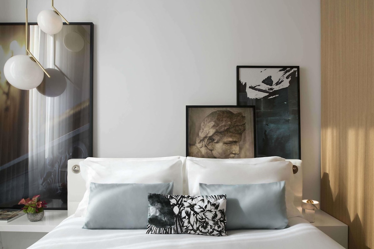 A modern bedroom with a bed, two pillows, a nightstand, a vase with flowers, and wall art.