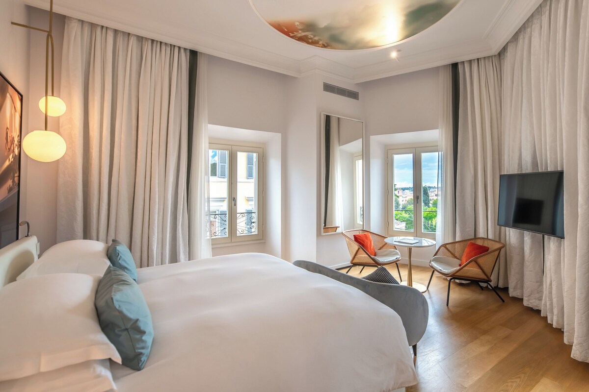 A hotel room with a large bed, a TV, a balcony view, and a dining area with chairs.
