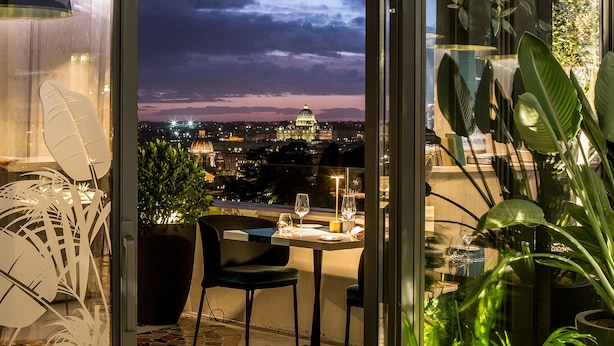 An open balcony with a table set for two, a potted plant, and a view of a cityscape at dusk. - Sofitel Rome Villa Borghese (Rome)