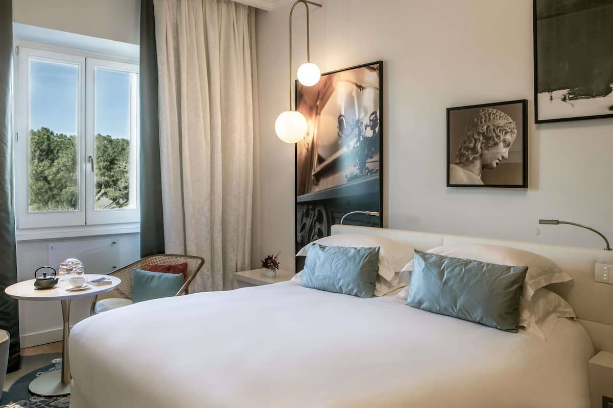 A hotel room with a large bed, a small round table with a kettle, a vase with flowers, and a window with curtains.