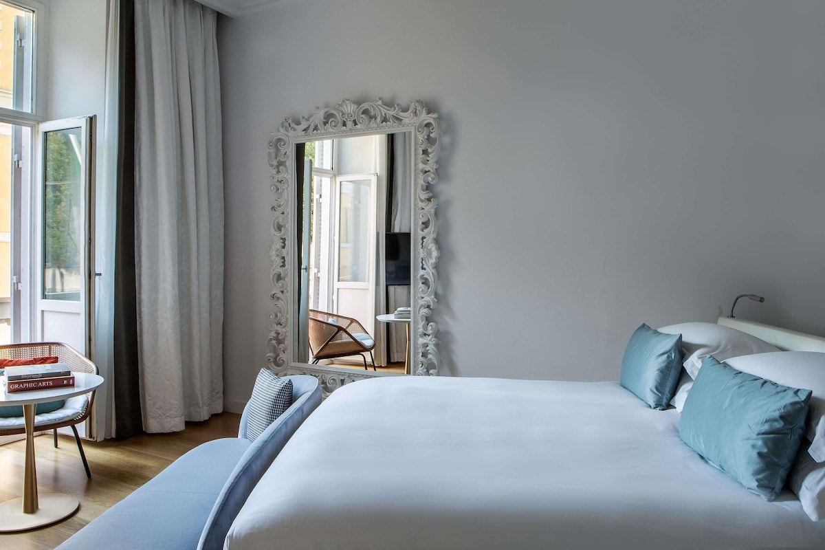 A hotel room with a large bed, a small table with books, a chair, and a large mirror.