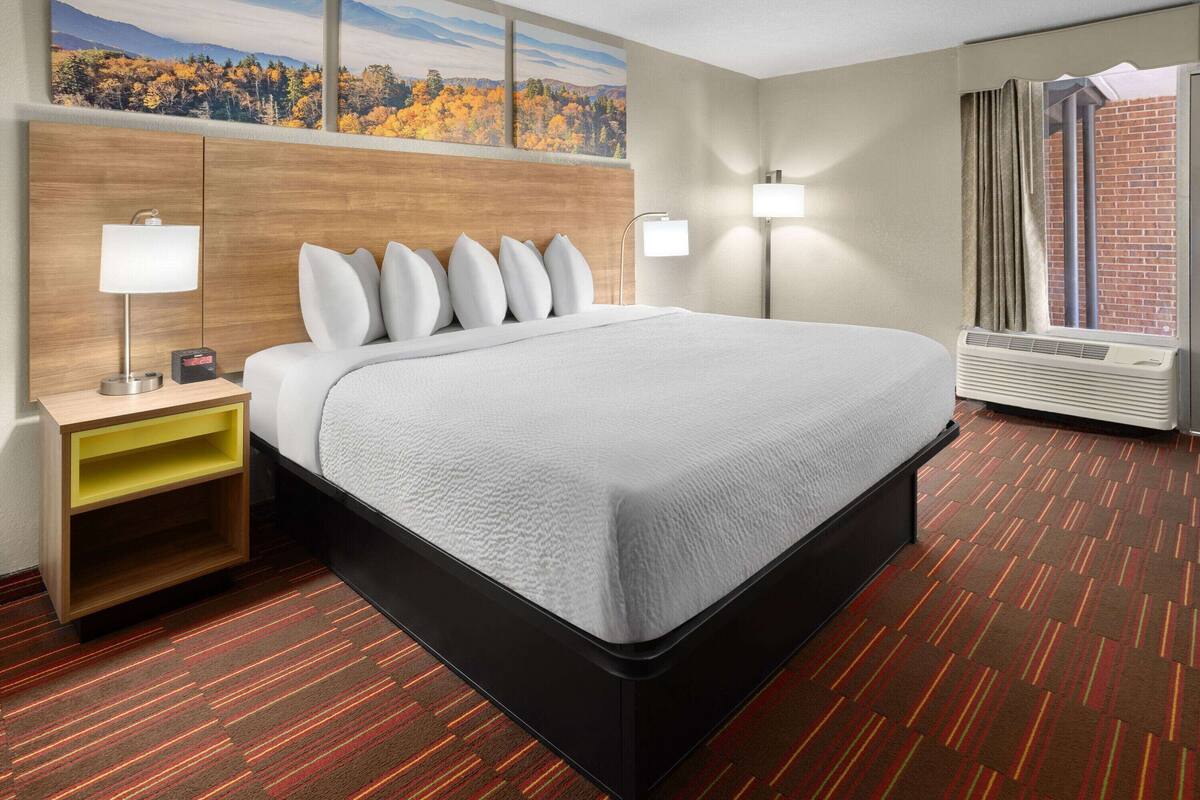 A hotel room with a large bed, two bedside tables, a wall-mounted lamp, a window with curtains, and a view of autumn foliage.
