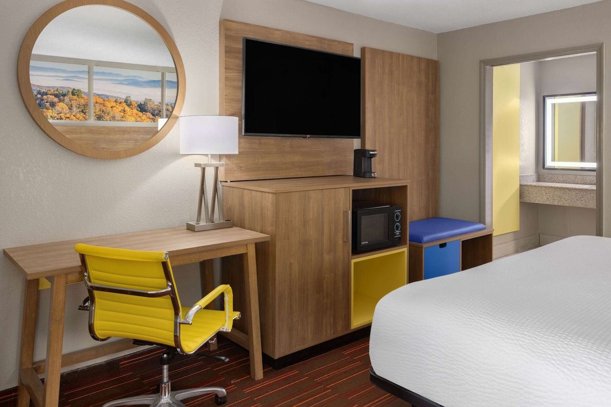 A hotel room with a bed, a desk with a chair, a TV, a coffee maker, a microwave, and a window with a view of autumn trees.