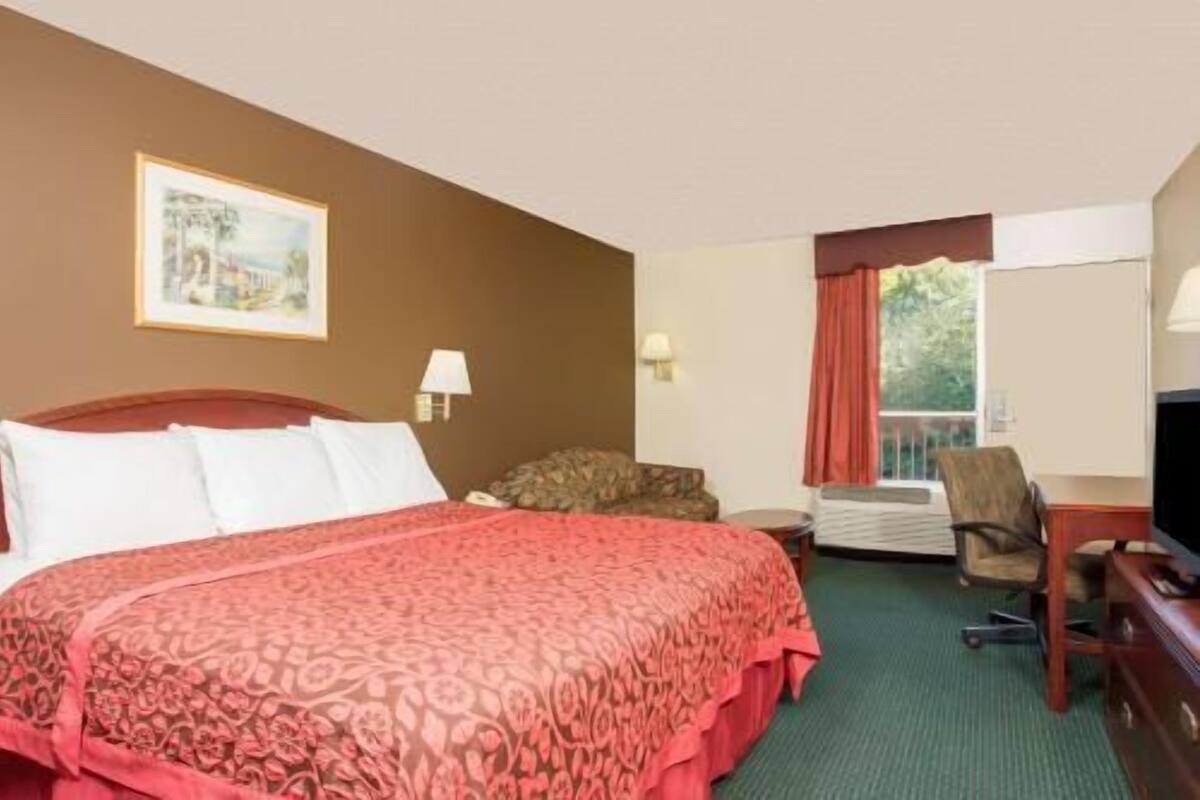 A hotel room with a bed, a chair, a desk, a lamp, and a window with curtains.