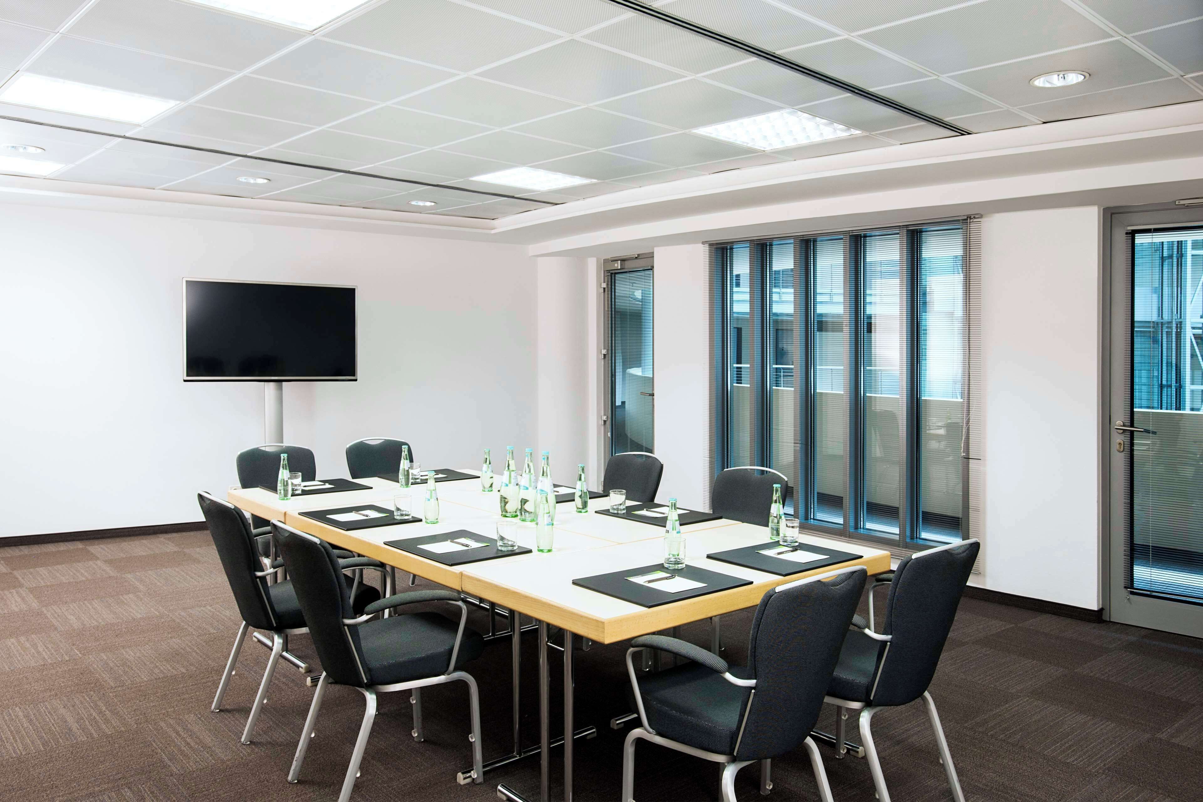 Un salón de conferencias con una mesa larga, sillas, un televisor de pantalla plana y una ventana con persianas.
