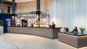 A reception desk in a modern hotel lobby with a large window, a reception area, and a staircase in the background. - NH Düsseldorf City (Düsseldorf)