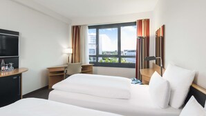 A hotel room with two beds, a desk, a TV, and a window with curtains. - NH Düsseldorf City (Düsseldorf)