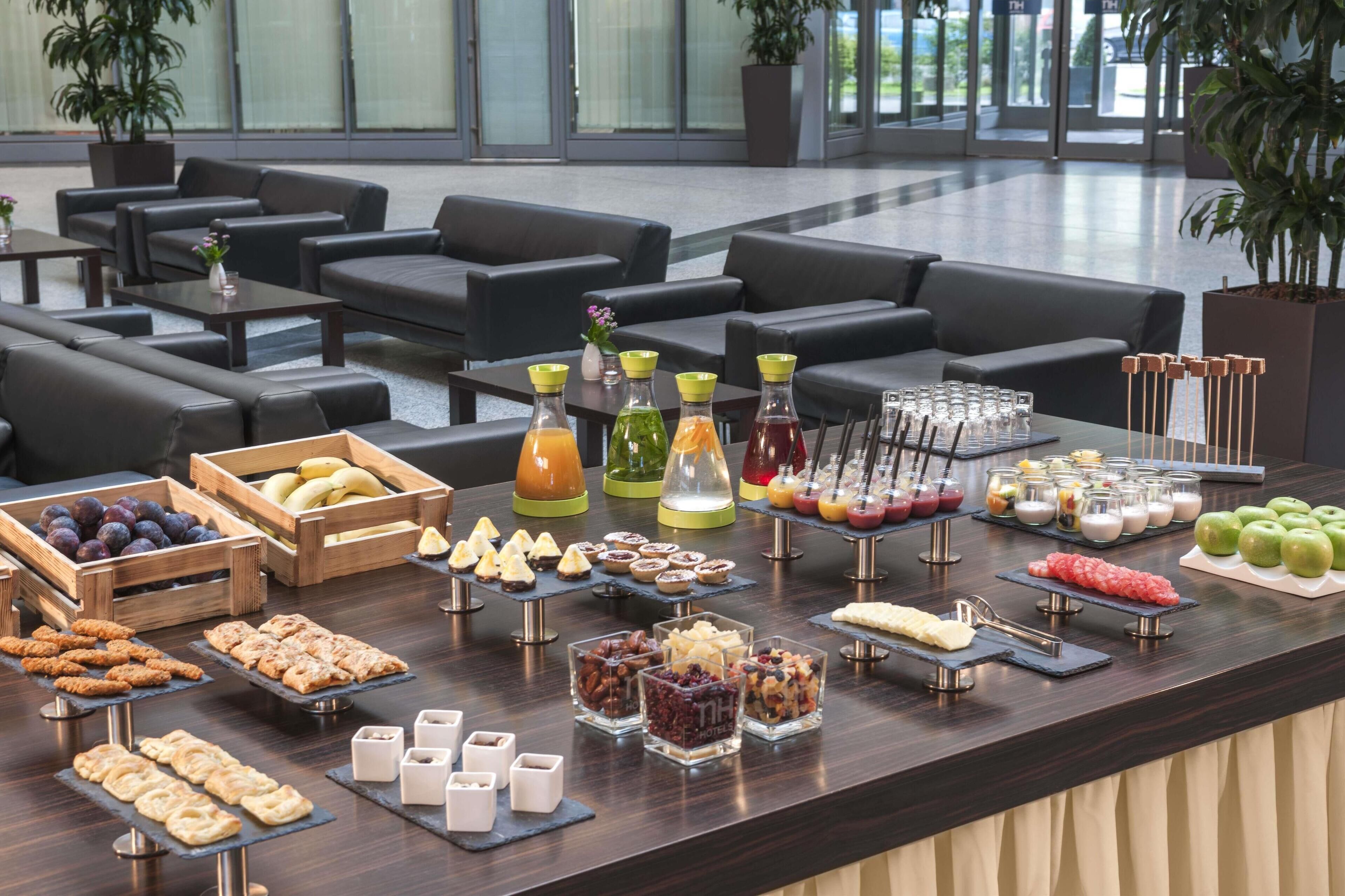 A buffet setup with various food items and beverages, including fruits, pastries, and drinks, in a modern indoor setting with seating areas.