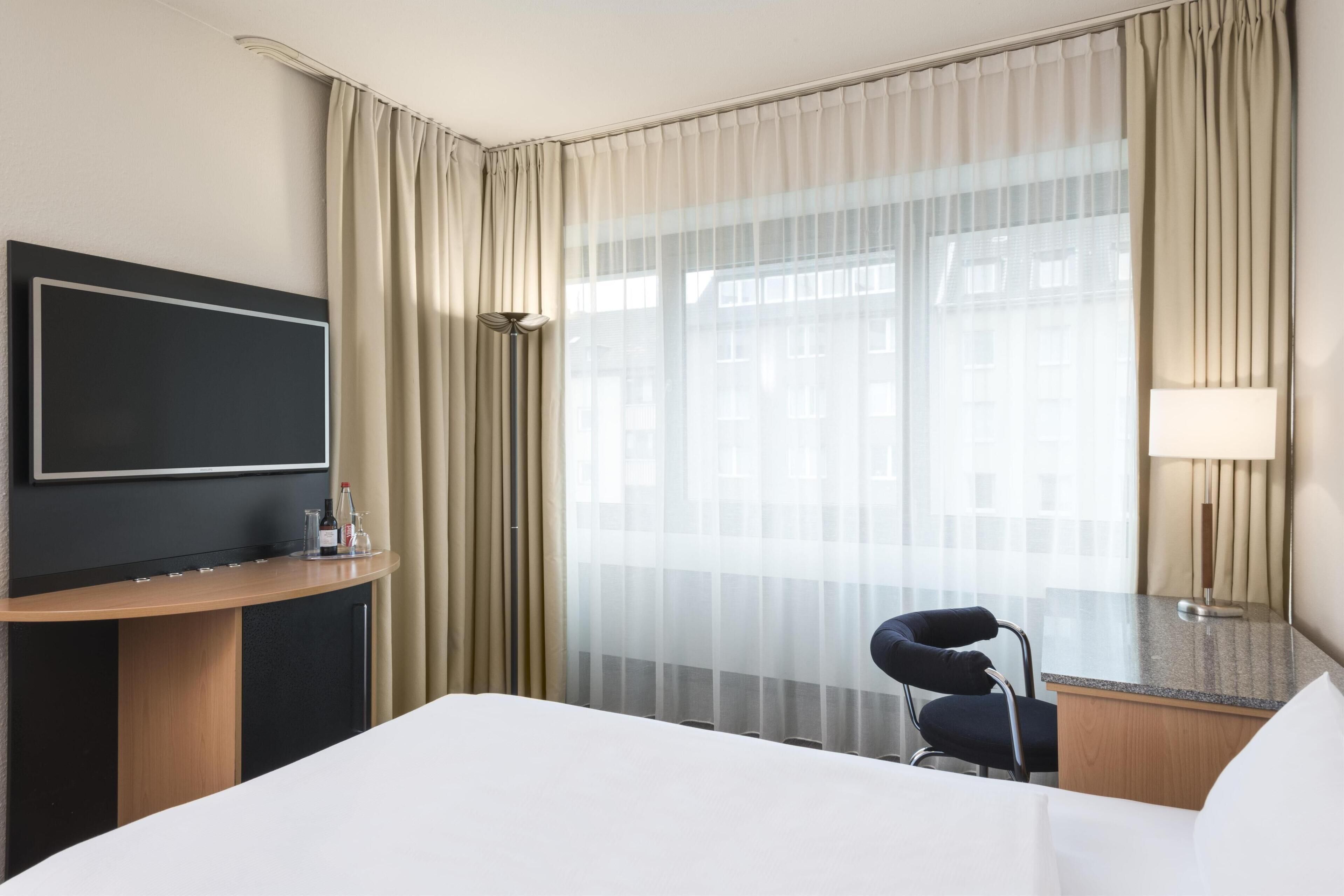 Una habitación de hotel con cama, escritorio con televisor, silla y ventana con cortinas.