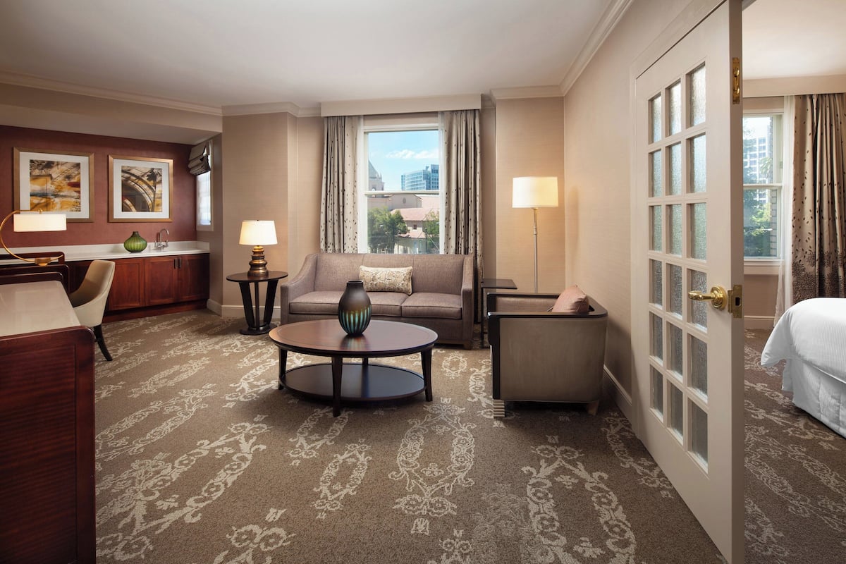 Ein Hotelzimmer mit einer Couch, einem Sessel und einem runden Tisch. Es gibt ein Fenster mit Vorhängen, eine Tür und einen Blick auf Gebäude.