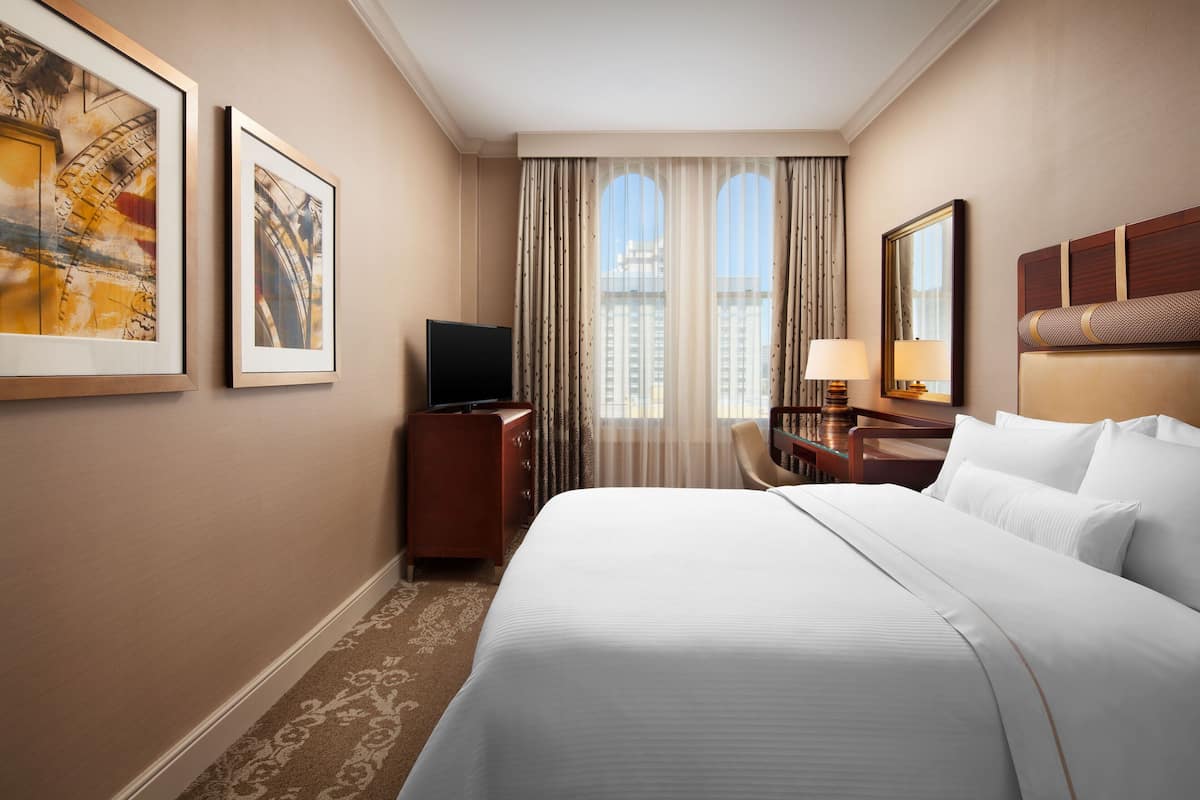Ein Hotelzimmer mit einem großen Bett, einem Schreibtisch mit Lampe, einem Fernseher und zwei gerahmten Kunstwerken an der Wand.