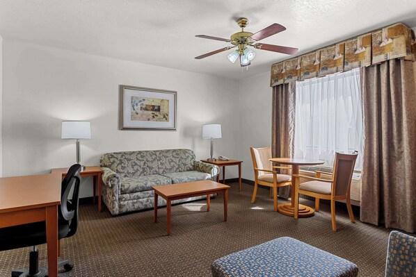 Une chambre d’hôtel avec un canapé, une petite table, un bureau, une chaise, un ventilateur de plafond et une fenêtre avec des rideaux.