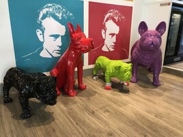 Una habitación con piso de madera, una estatua de perro negro y coloridas estatuas de animales frente a una pared con un mural rojo y azul.