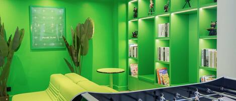 Una habitación con decoración en tonos verdes, una mesa de fútbol de mesa, un sofá, una pequeña mesa redonda y estanterías con libros y objetos decorativos.