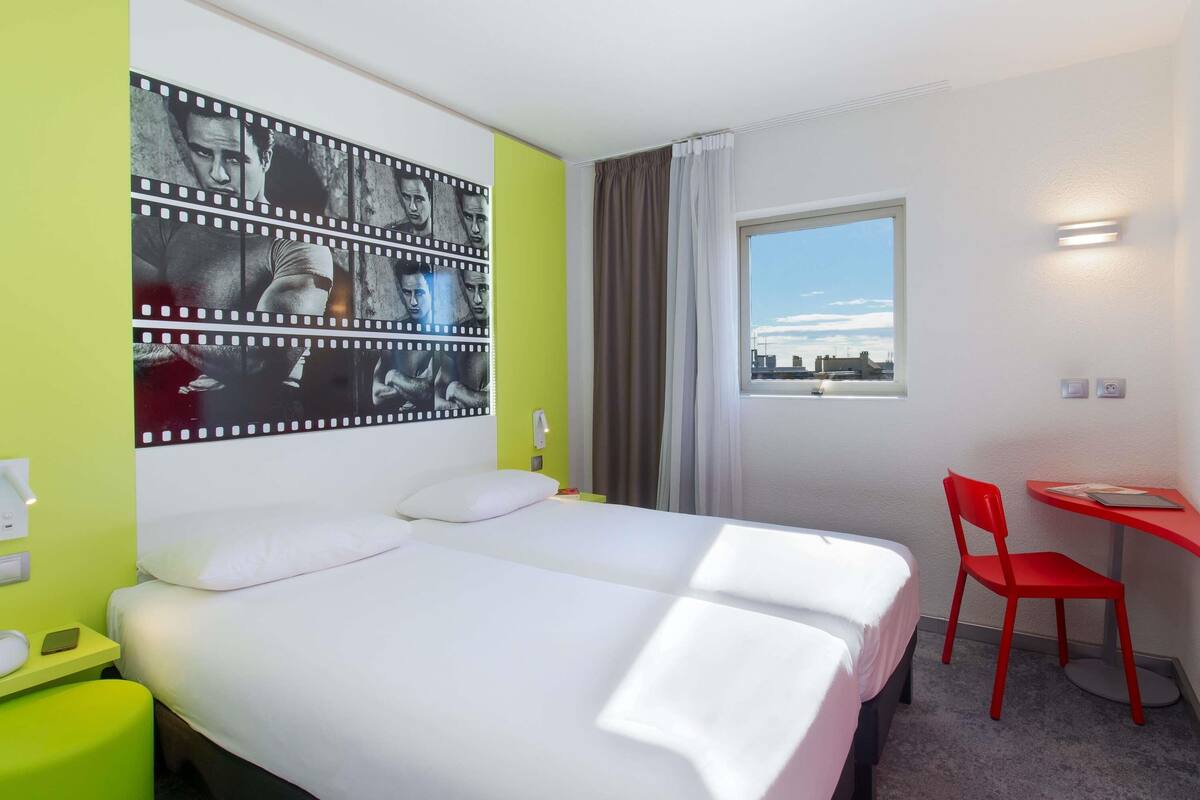 Quarto de hotel com uma cama, uma escrivaninha com cadeira, janela com vista e uma obra de arte com tema de filme na parede.