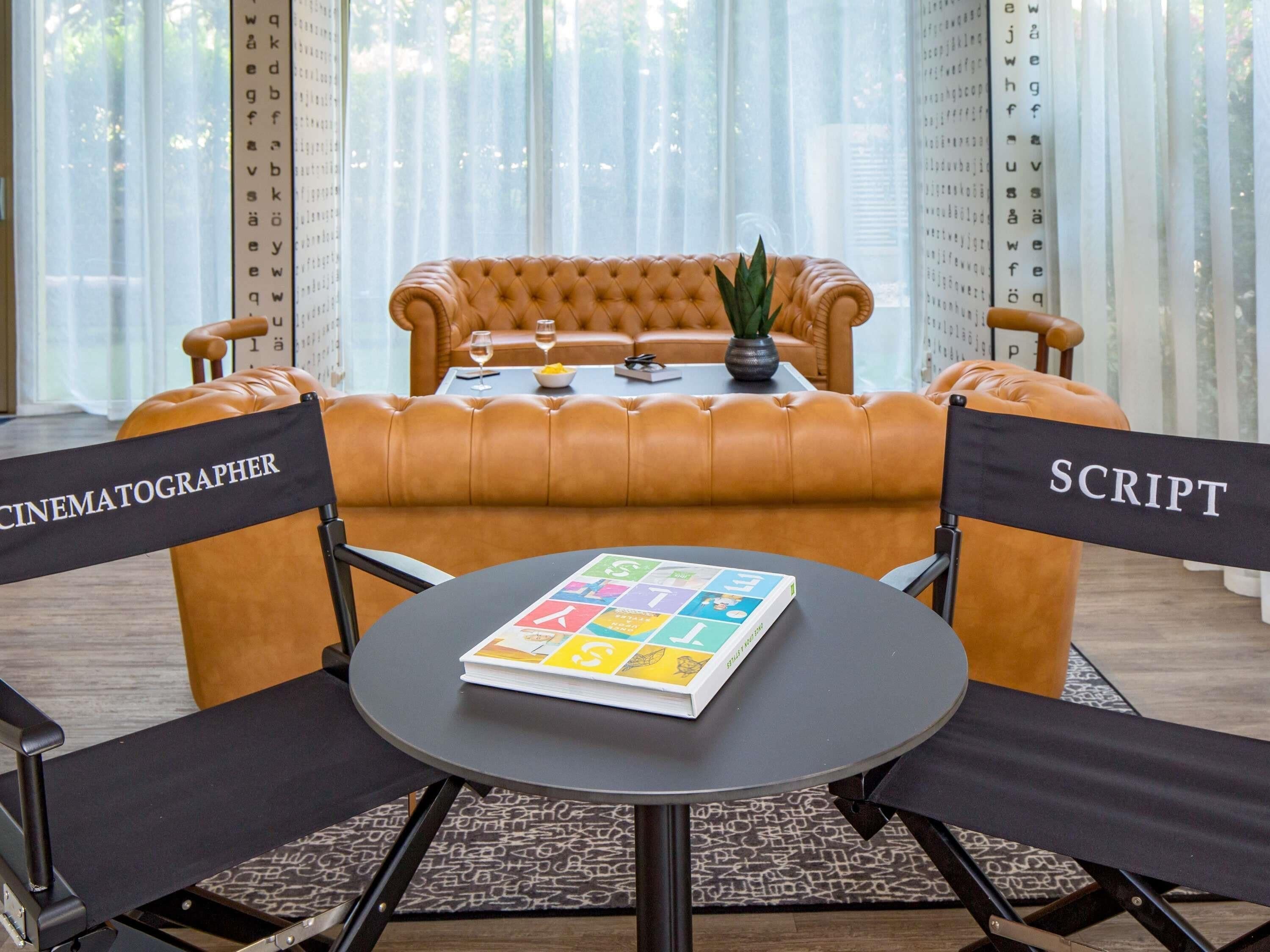 Una habitación con un sofá de cuero, una mesa con un libro y dos sillas con letreros que dicen 'CINEMATOGRAPHER' y 'SCRIPT'.