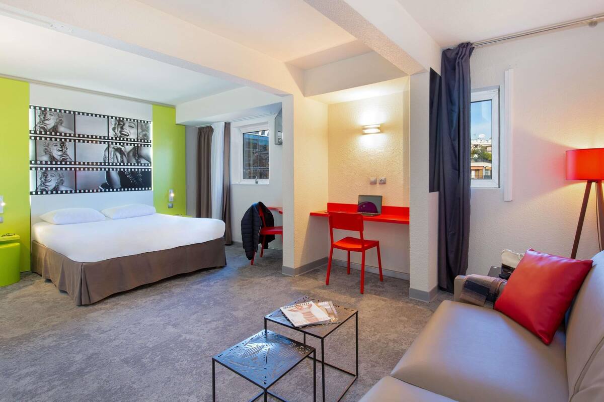 Quarto de hotel com uma cama, uma escrivaninha com cadeira, um sofá, uma mesinha e um painel decorado com uma faixa de filme.
