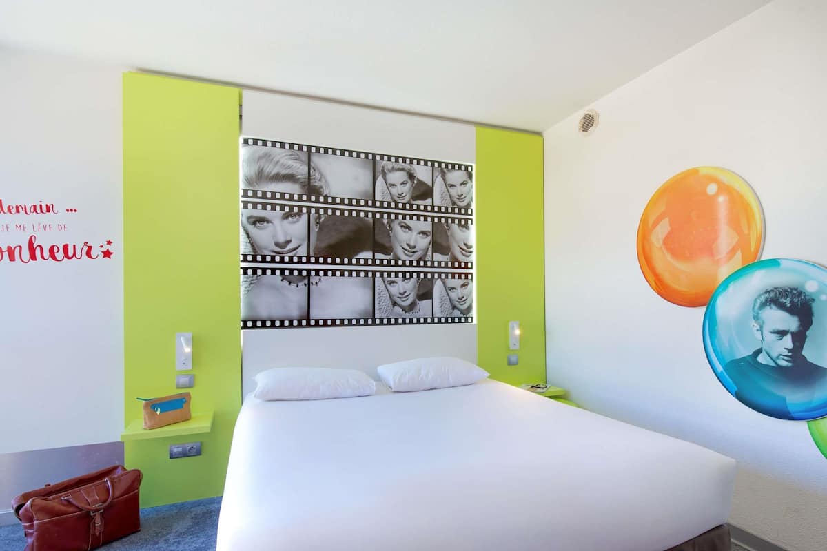 Quarto de hotel com cama, cabeceira com colagem de fotos em estilo de fita de filme, parede verde e mesa de cabeceira com abajur.