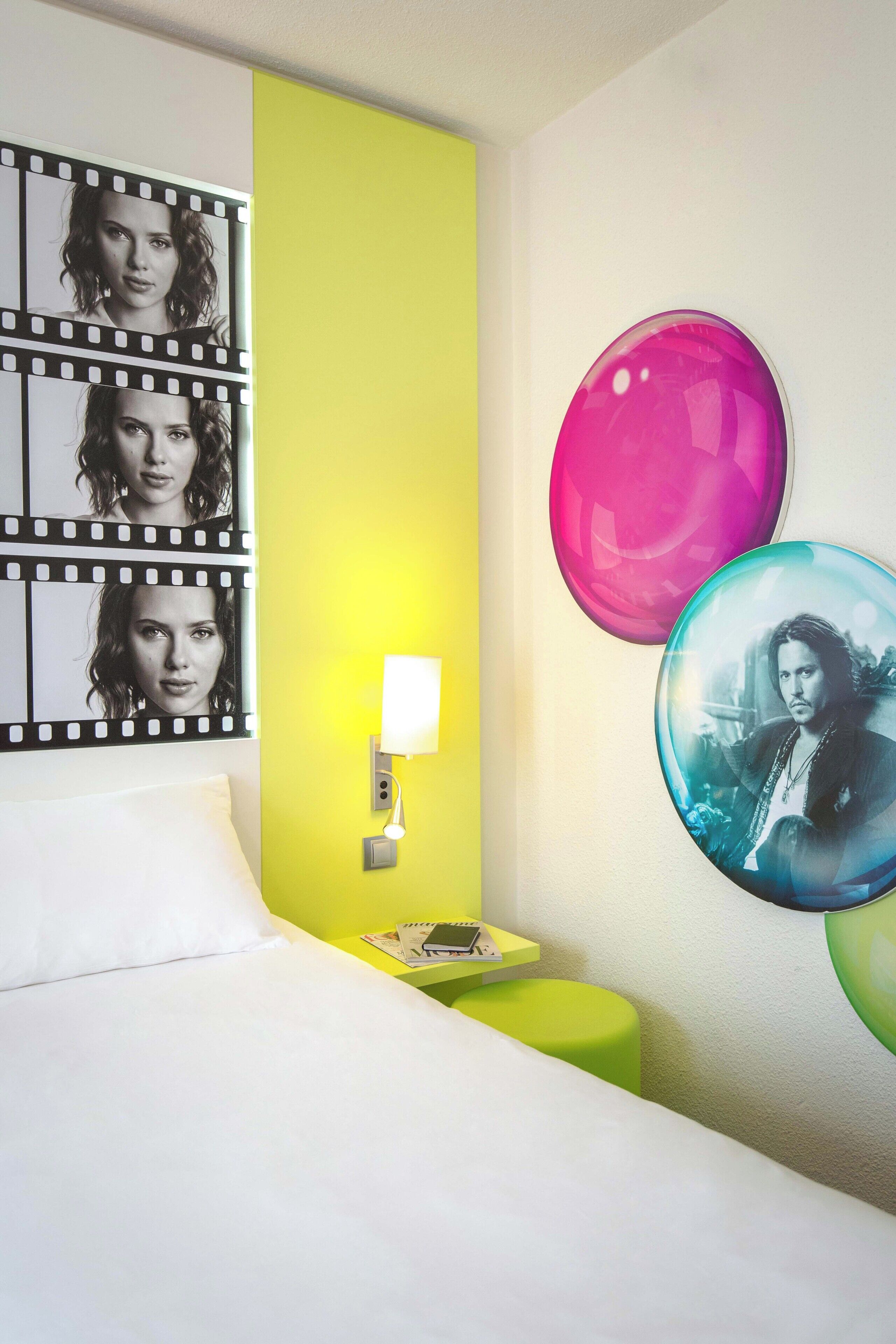 Een hotelkamer met een bed, nachtkastje, lamp, groene muur en een stripkunstwerk.