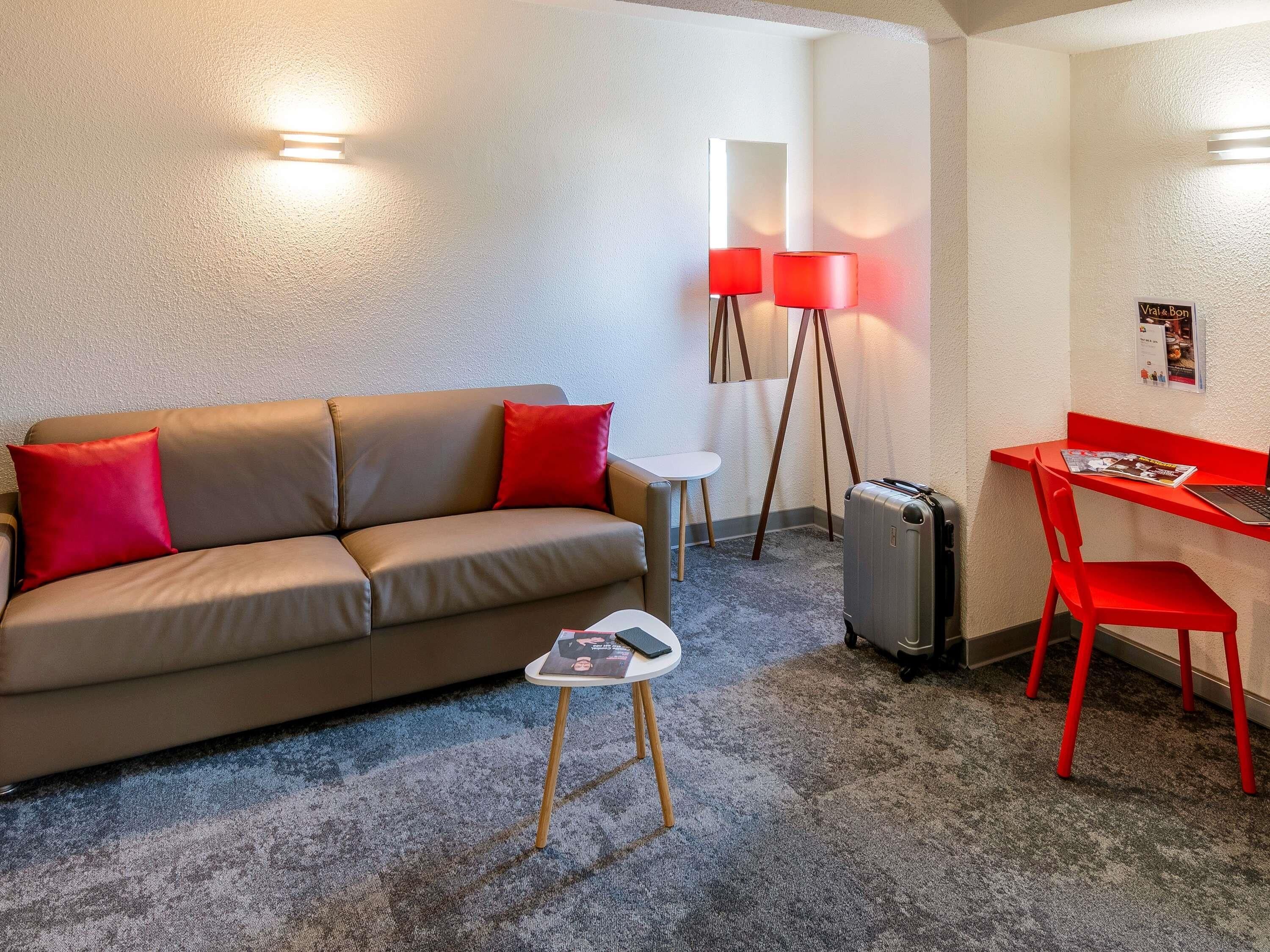 Een kamer met een bruine bank, rode kussens, een tafeltje met een tijdschrift, een koffer, een rode stoel, een bureau met een laptop en twee vloerlampen.