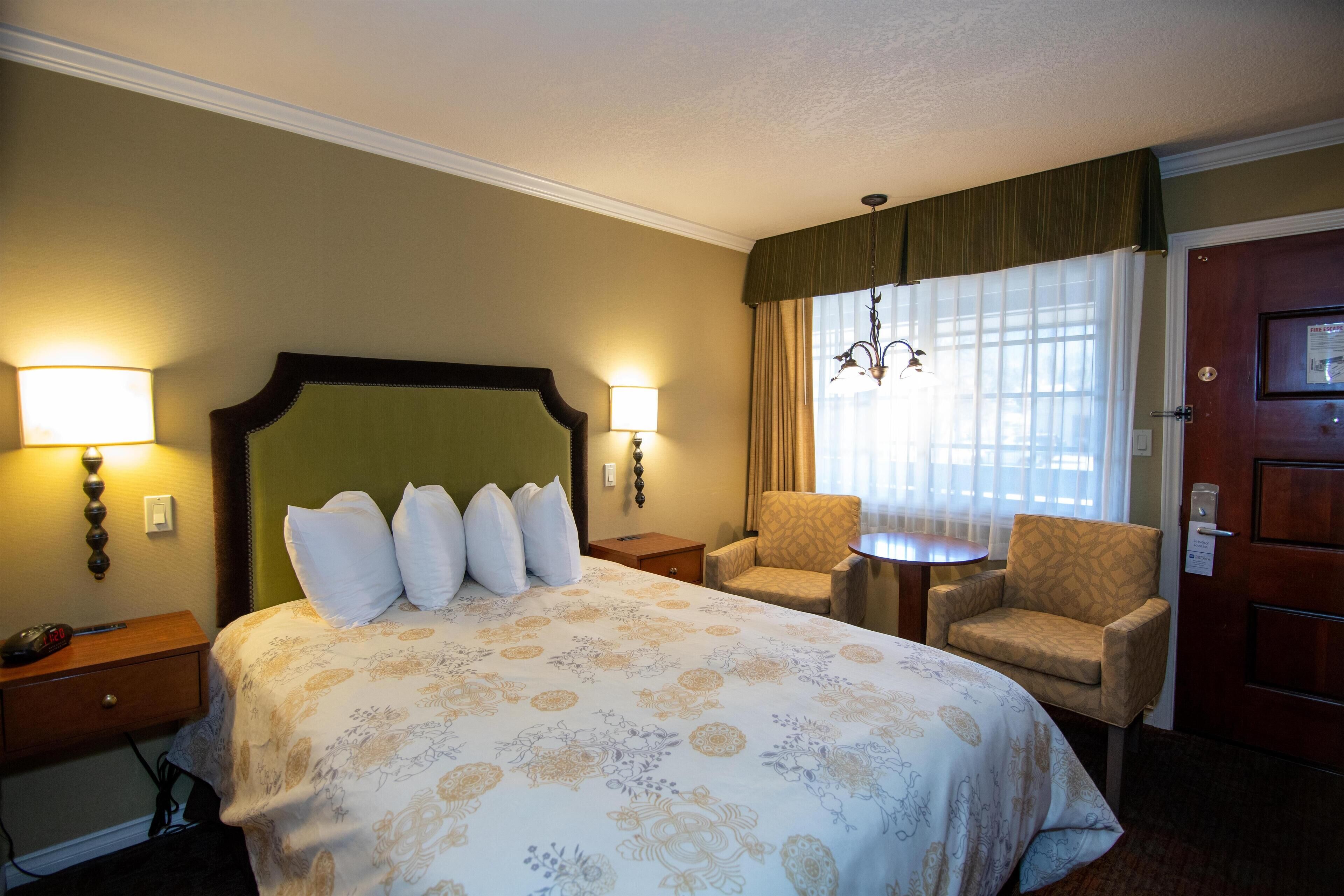 A hotel room with a large bed, two bedside lamps, a chair, a small table, and a door.