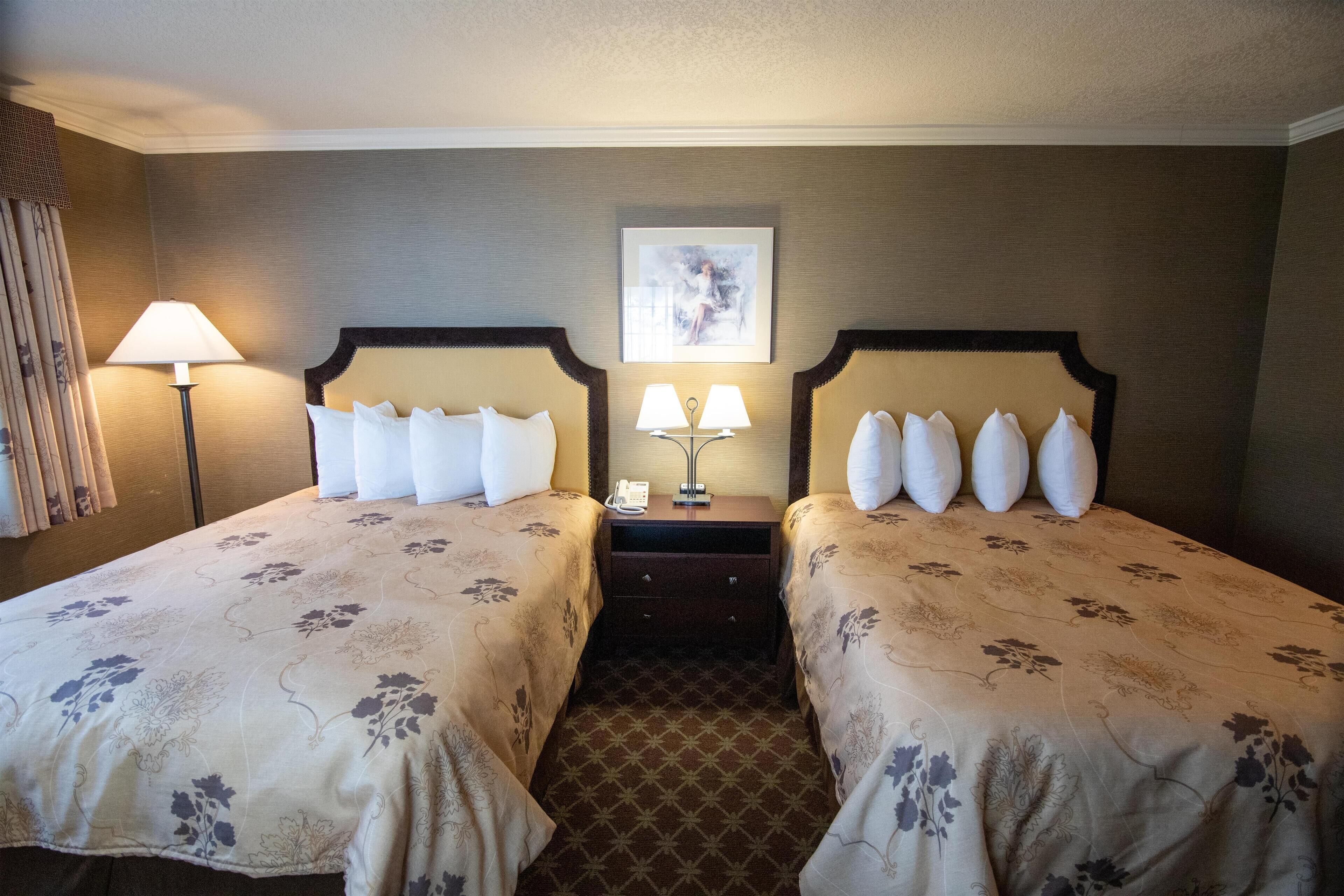 Habitación de hotel con dos camas, cada una con cubrecama floral y varios almohadones. Entre las camas, un mesita de noche con lámpara y teléfono. Encima de la mesita, un cuadro enmarcado en la pared.