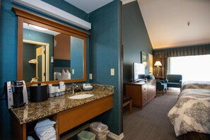 A hotel room with a granite countertop sink, a mirror, a desk with a lamp, a chair, and a bed with a patterned bedspread.