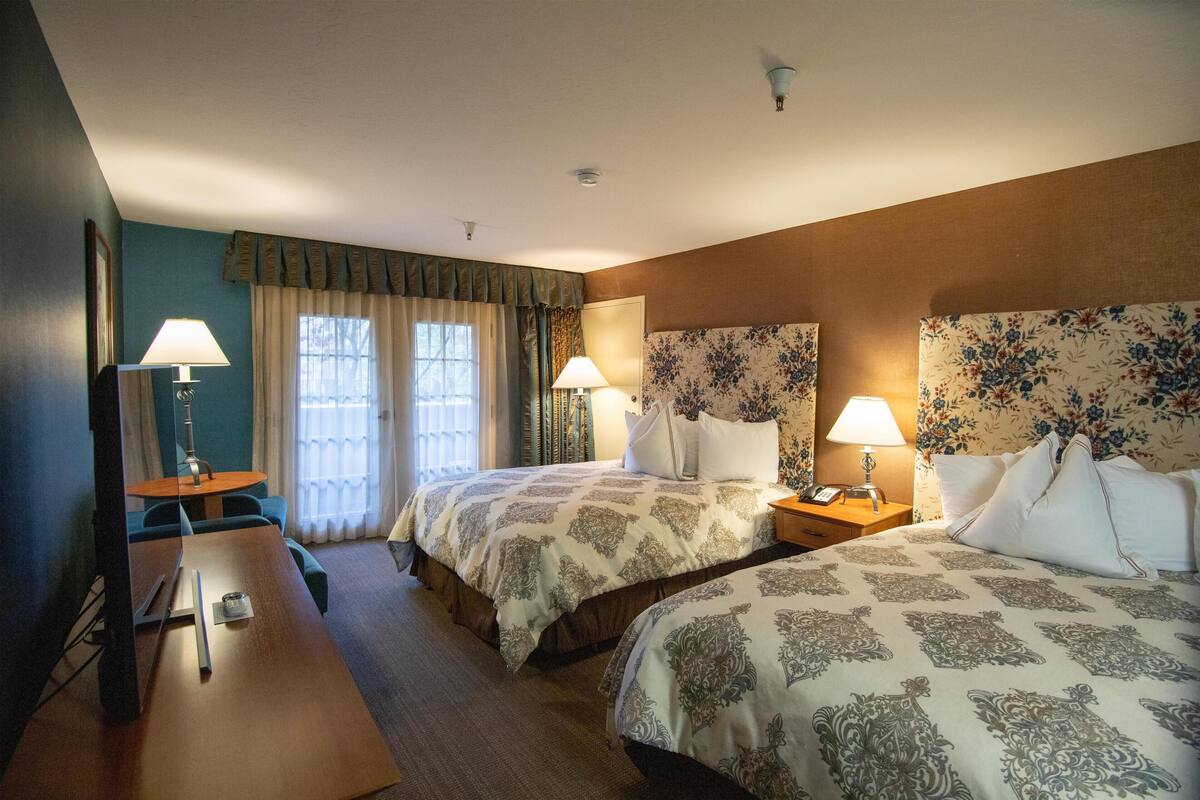 A hotel room with two beds, a desk, and a chair. There are lamps on each side of the beds and a table with a lamp.