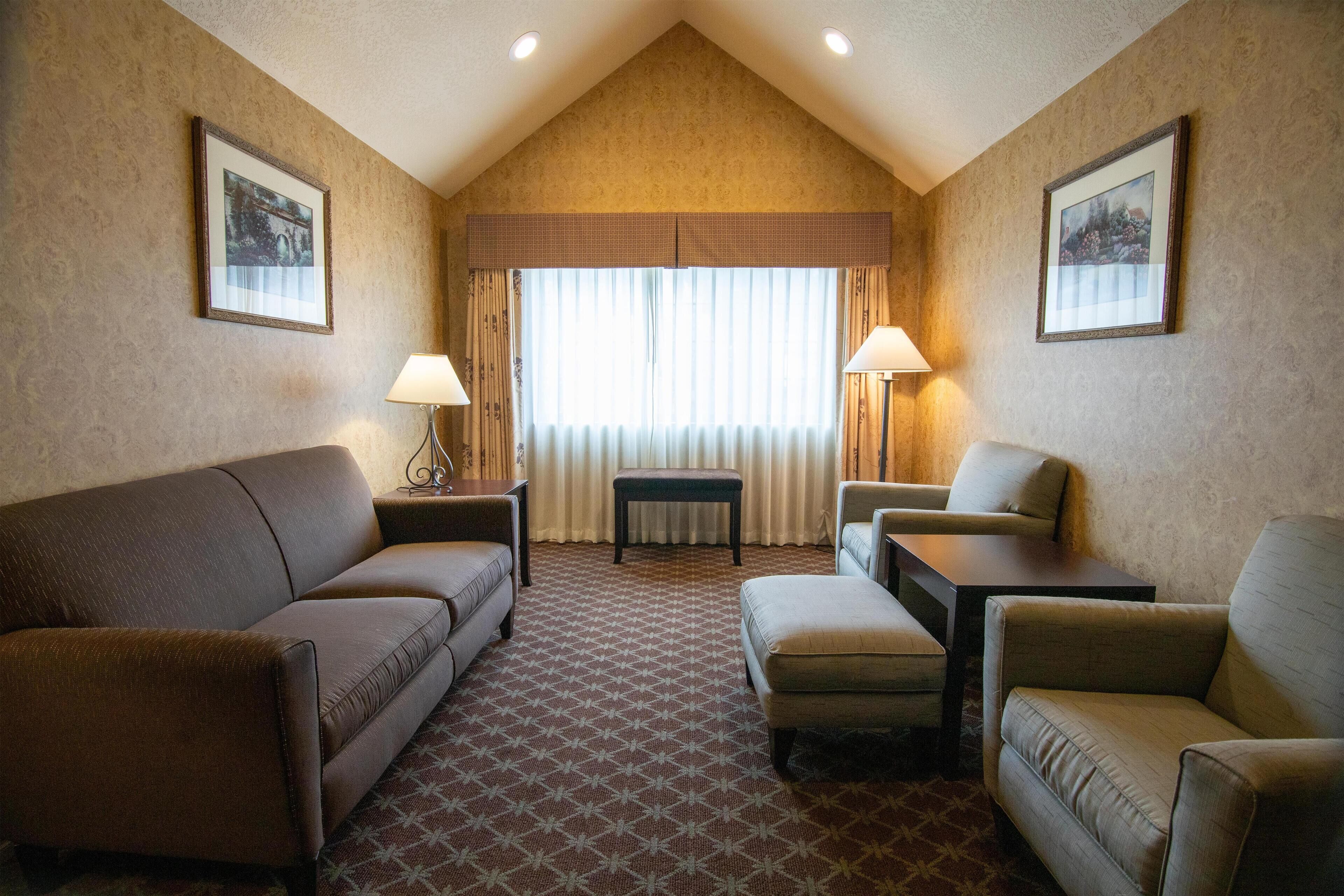 A hotel room with a brown patterned carpet, a brown sofa, a small table, a chair, and two lamps.