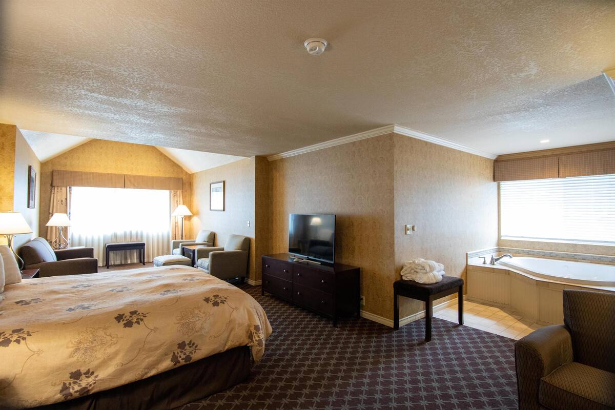 A hotel room with a large bed, a TV, a desk, a chair, a small table, and a bathroom with a bathtub.