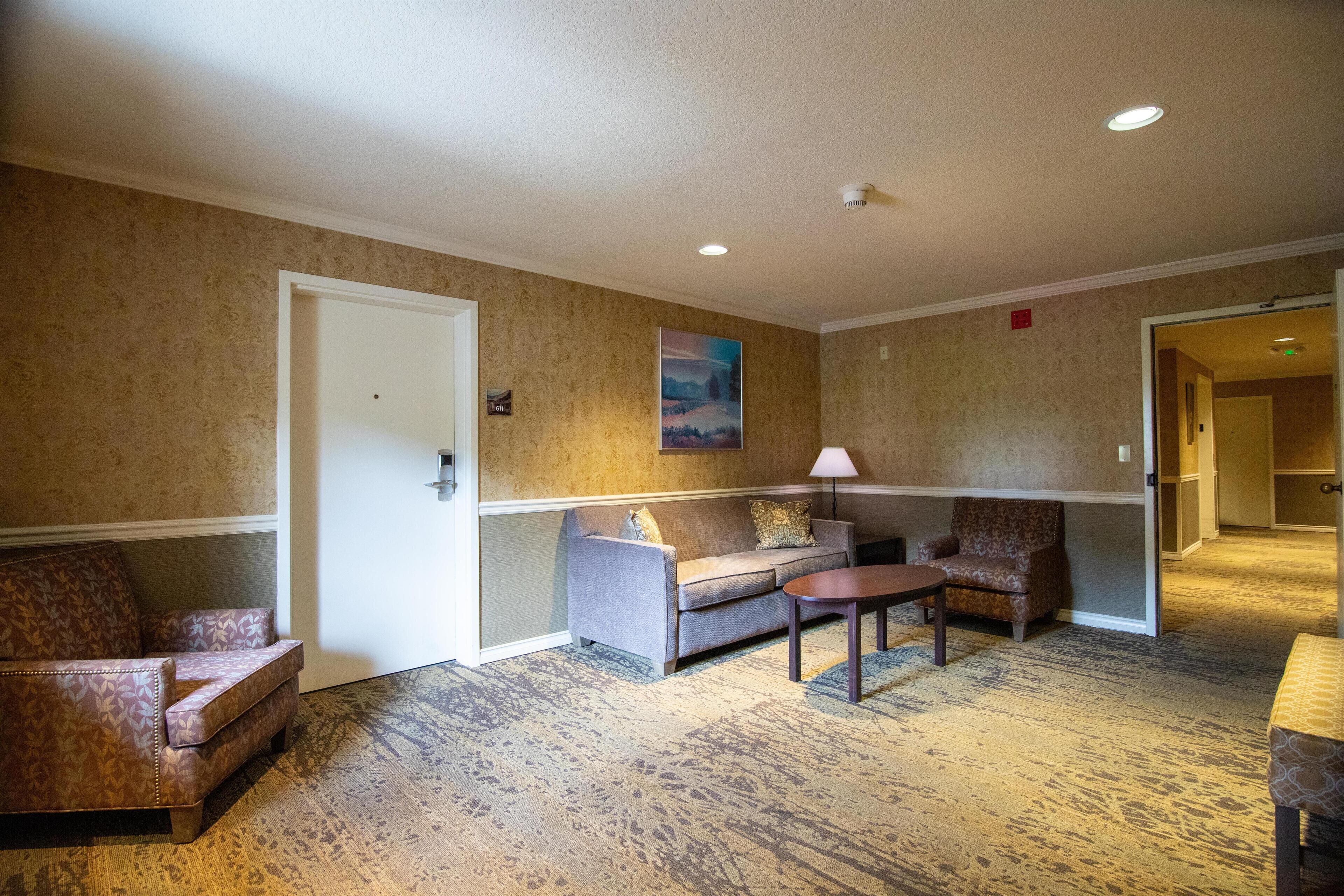 Um hall de hotel com carpete estampado, um sofá cinza, uma poltrona estampada e uma mesa pequena.