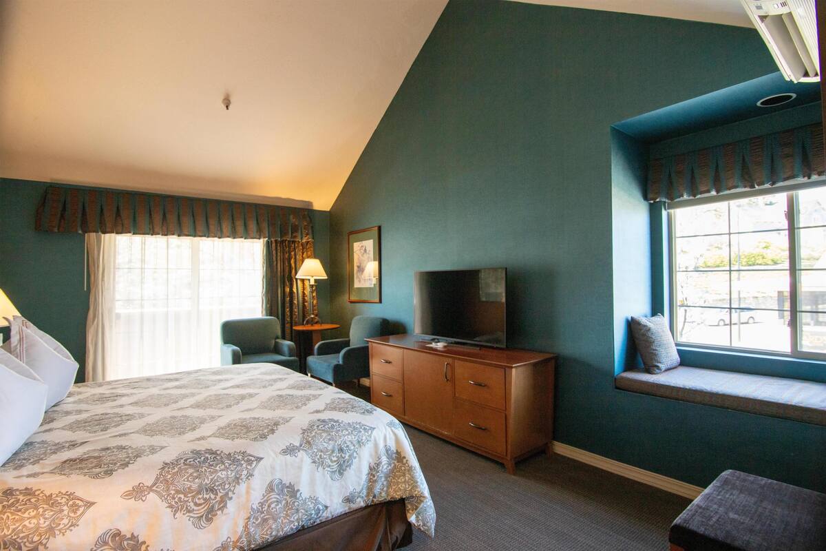 A bedroom with a bed, a dresser, a television, a window seat, and a desk with a lamp.