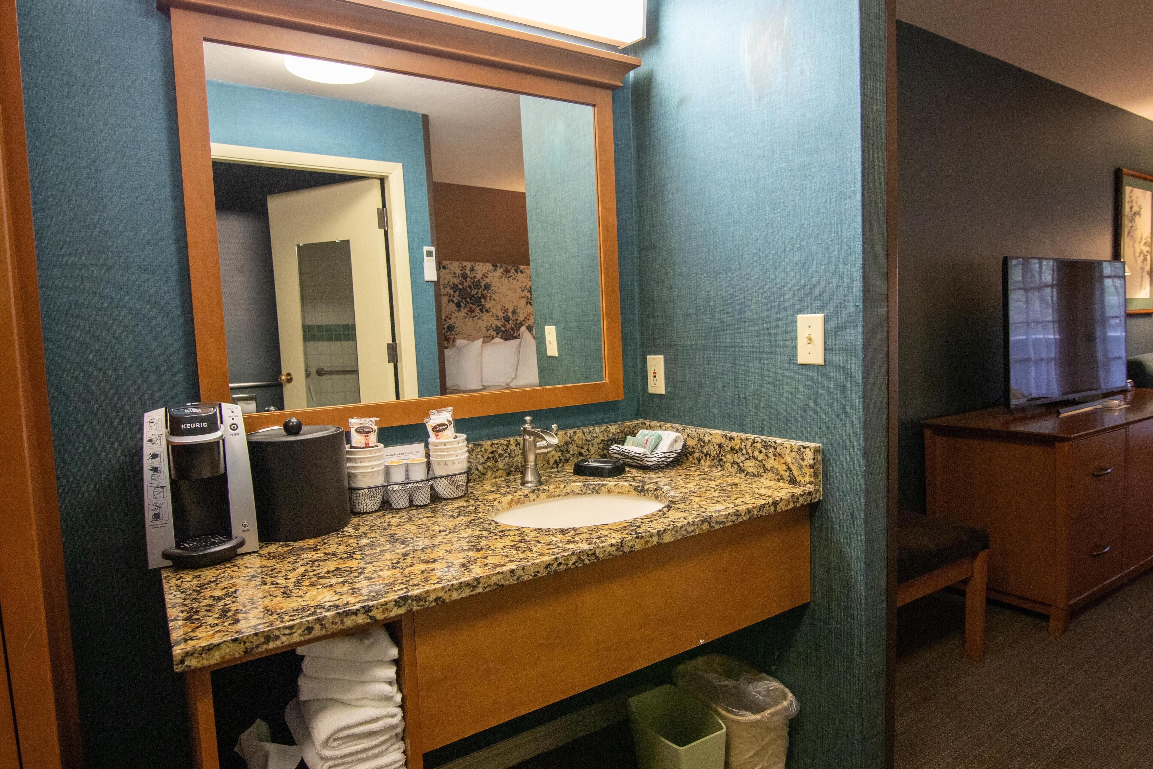 Une salle de bain d’hôtel équipée d’un comptoir en granit, d’une machine à café, d’un lavabo, d’un miroir, d’une télévision et d’un meuble de rangement.