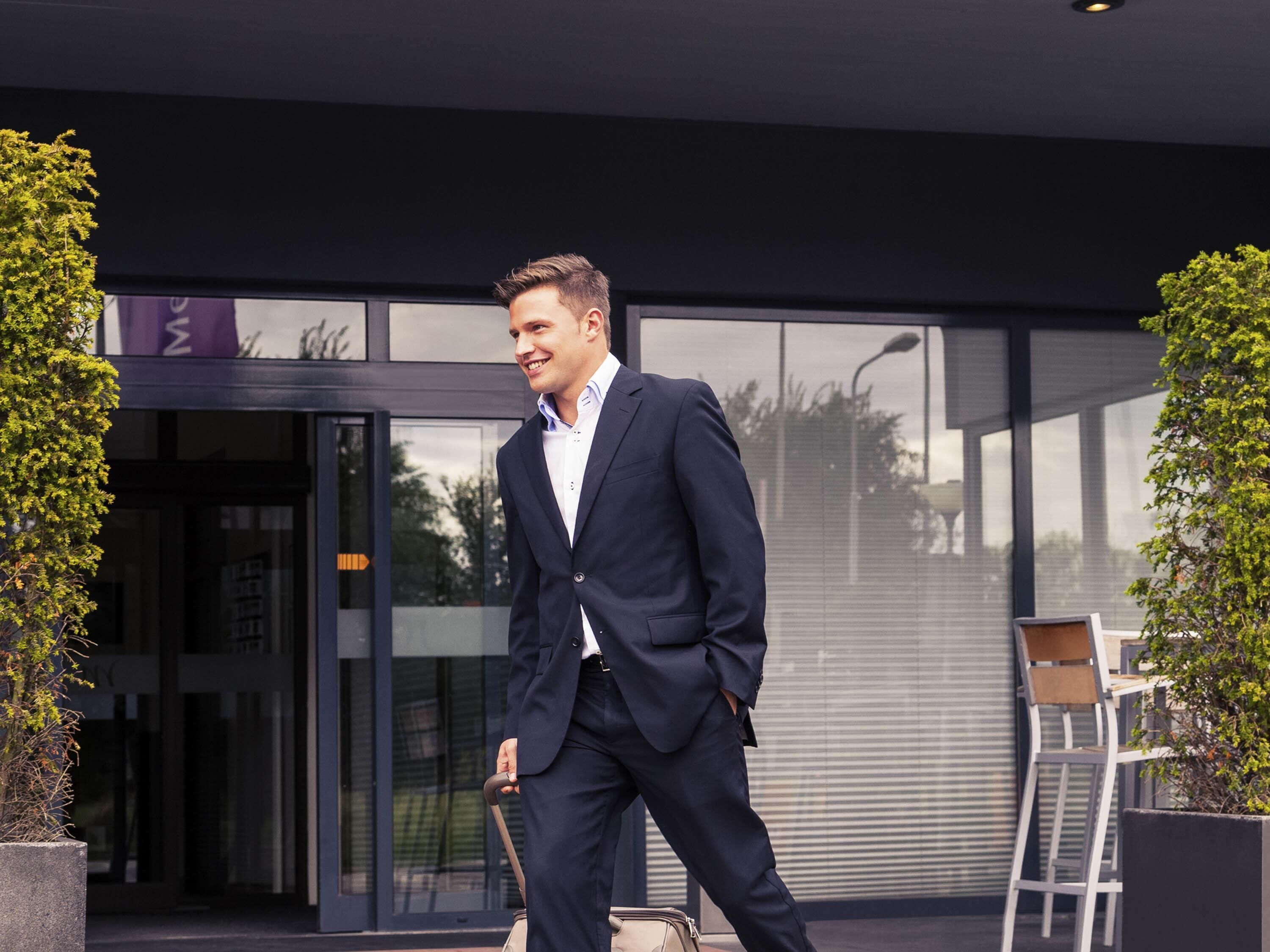Un hombre con traje caminando con una maleta, la entrada de un edificio moderno y mesas al aire libre.