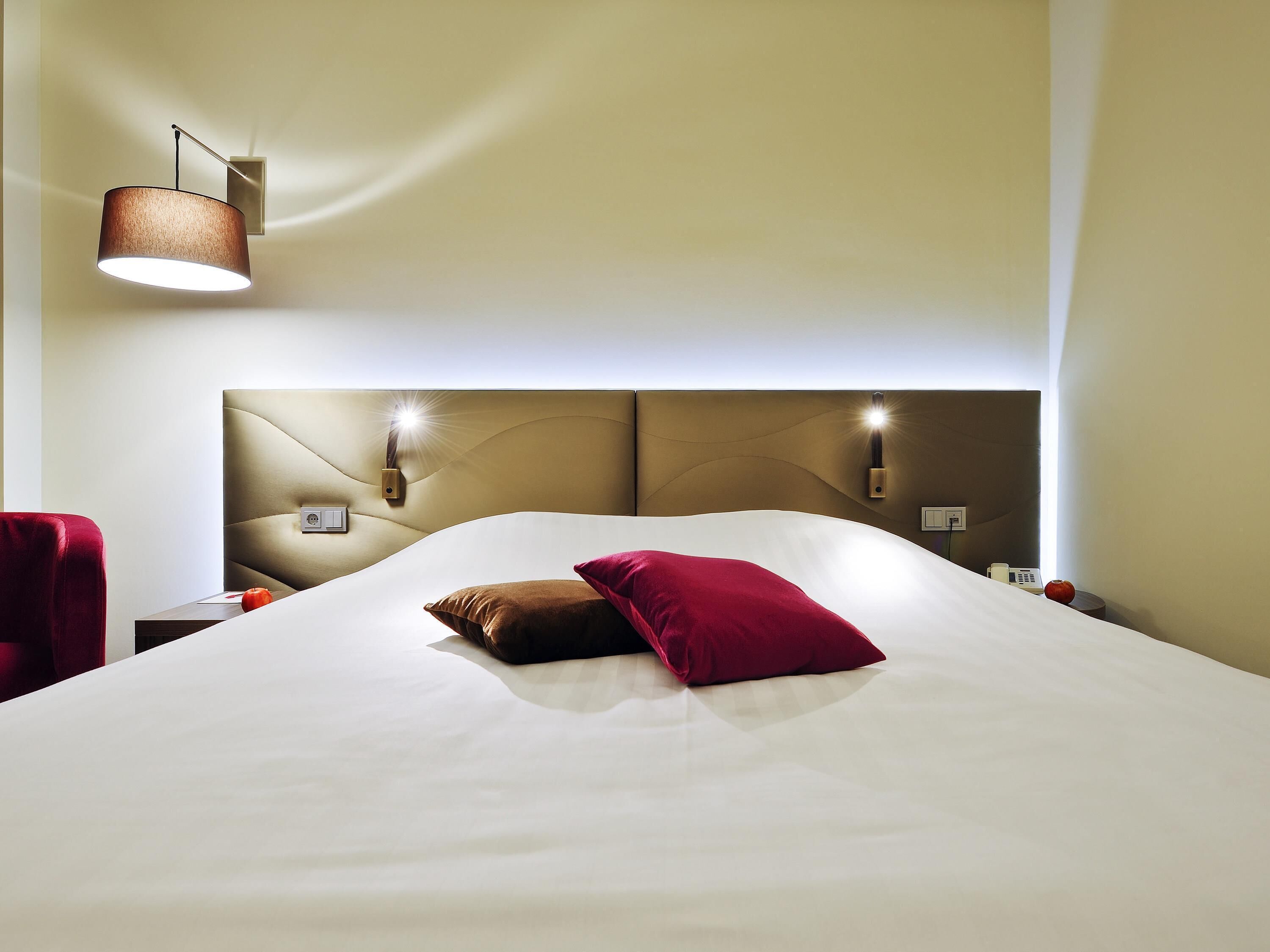 A hotel room with a large bed, a headboard with integrated lighting, a bedside table, and a chair.