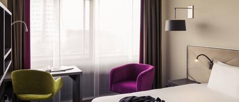 A hotel room with a bed, a desk, a chair, and a nightstand with a lamp.