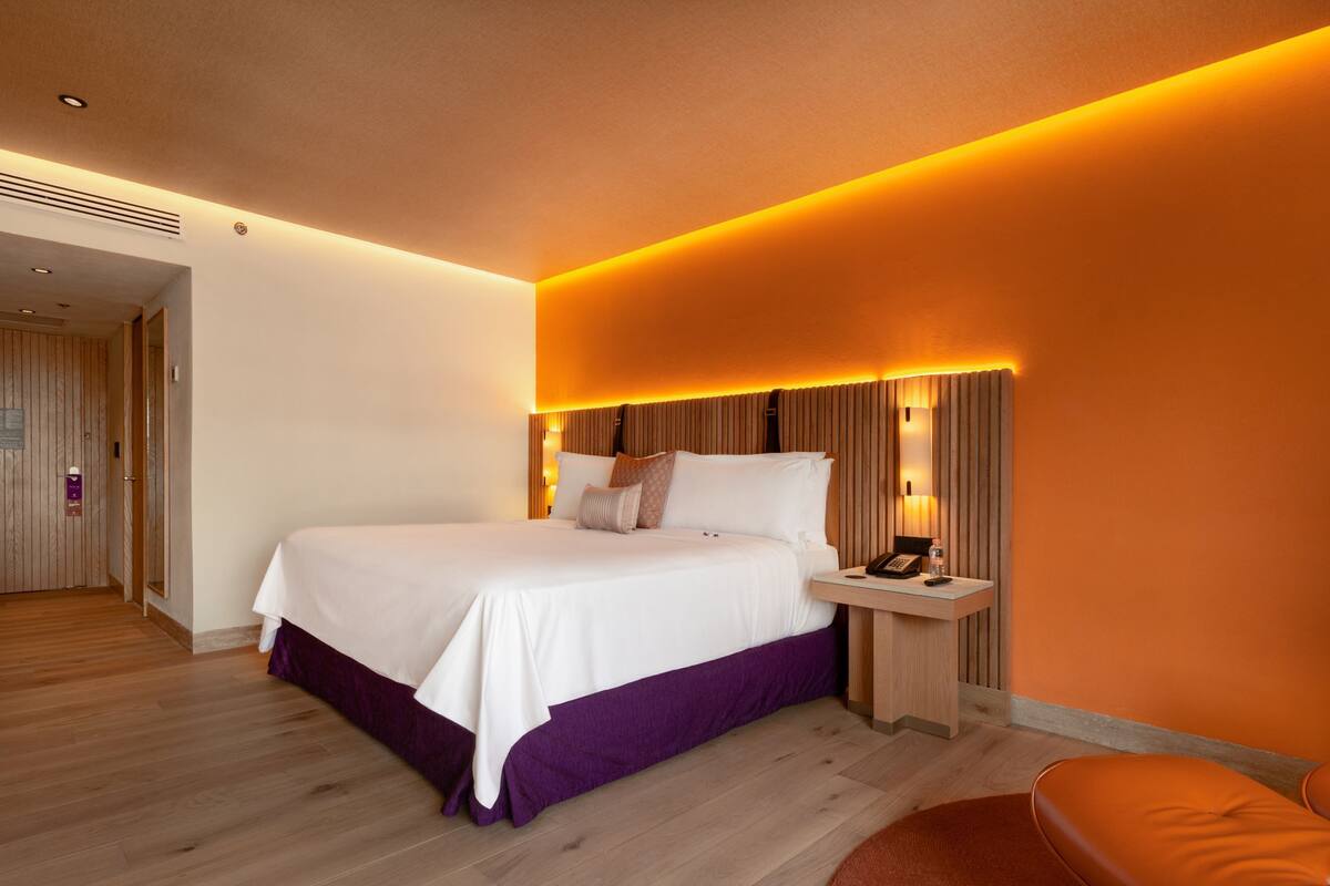 A modern hotel room with a large bed, a bedside table with a lamp, and a wooden floor.