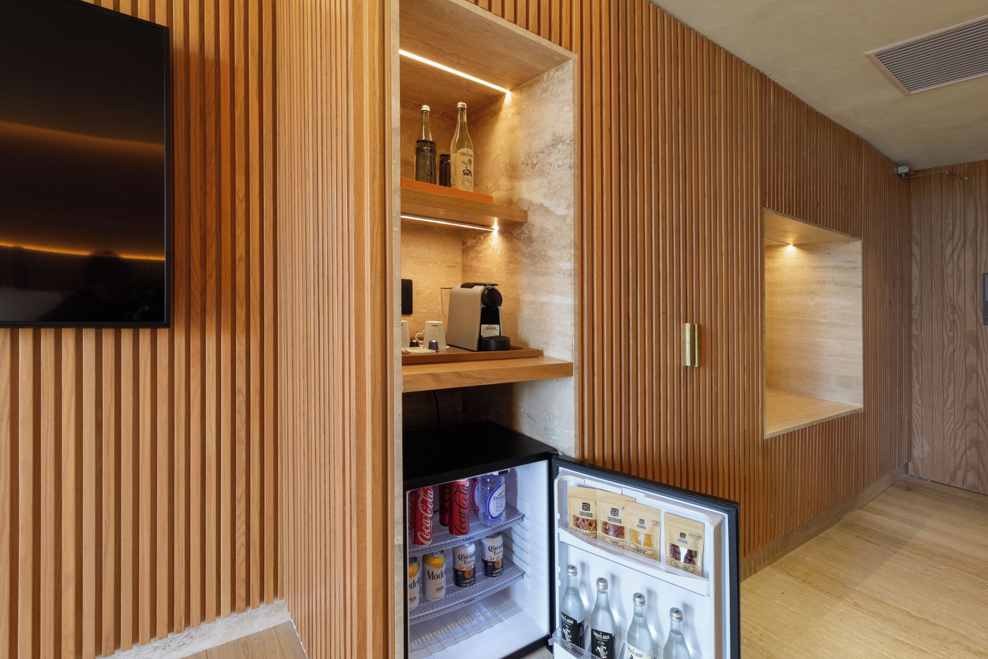 Quarto de hotel compacto com parede revestida em madeira, TV de tela plana na parede, minibar com bebidas e petiscos, e uma pequena geladeira.