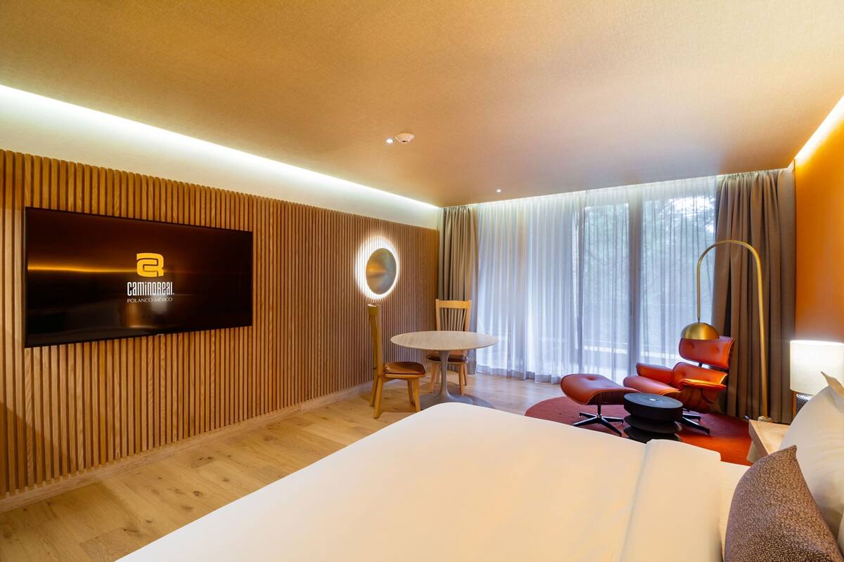 A modern hotel room with a large bed, a flat-screen TV mounted on the wall, a round mirror, a round table, and red chairs.