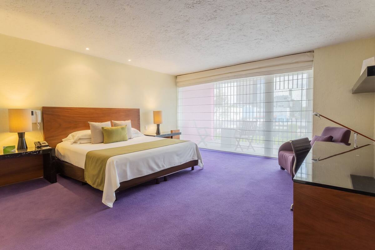 A hotel room with a large bed, a nightstand, a desk with a lamp, a chair, and a window with blinds.