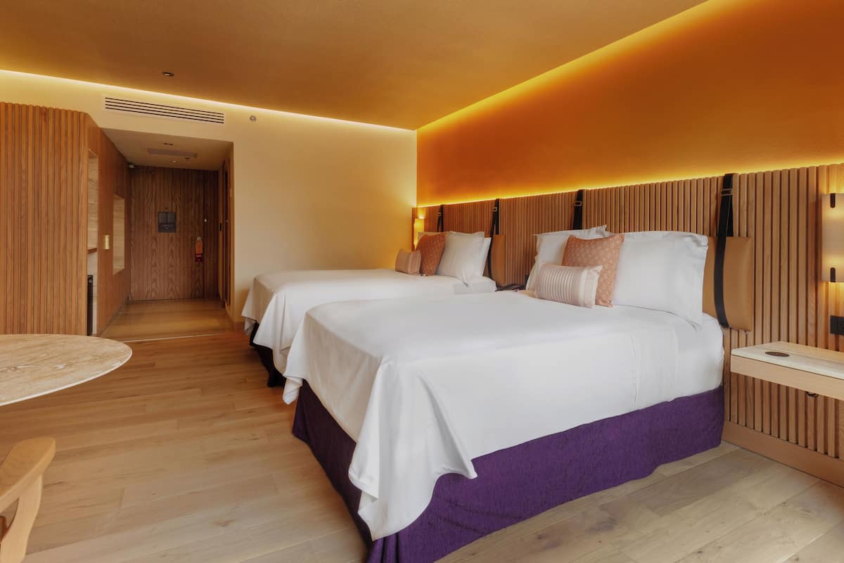A hotel room with two beds, a wooden floor, a small round table, and a wooden headboard with integrated lighting.