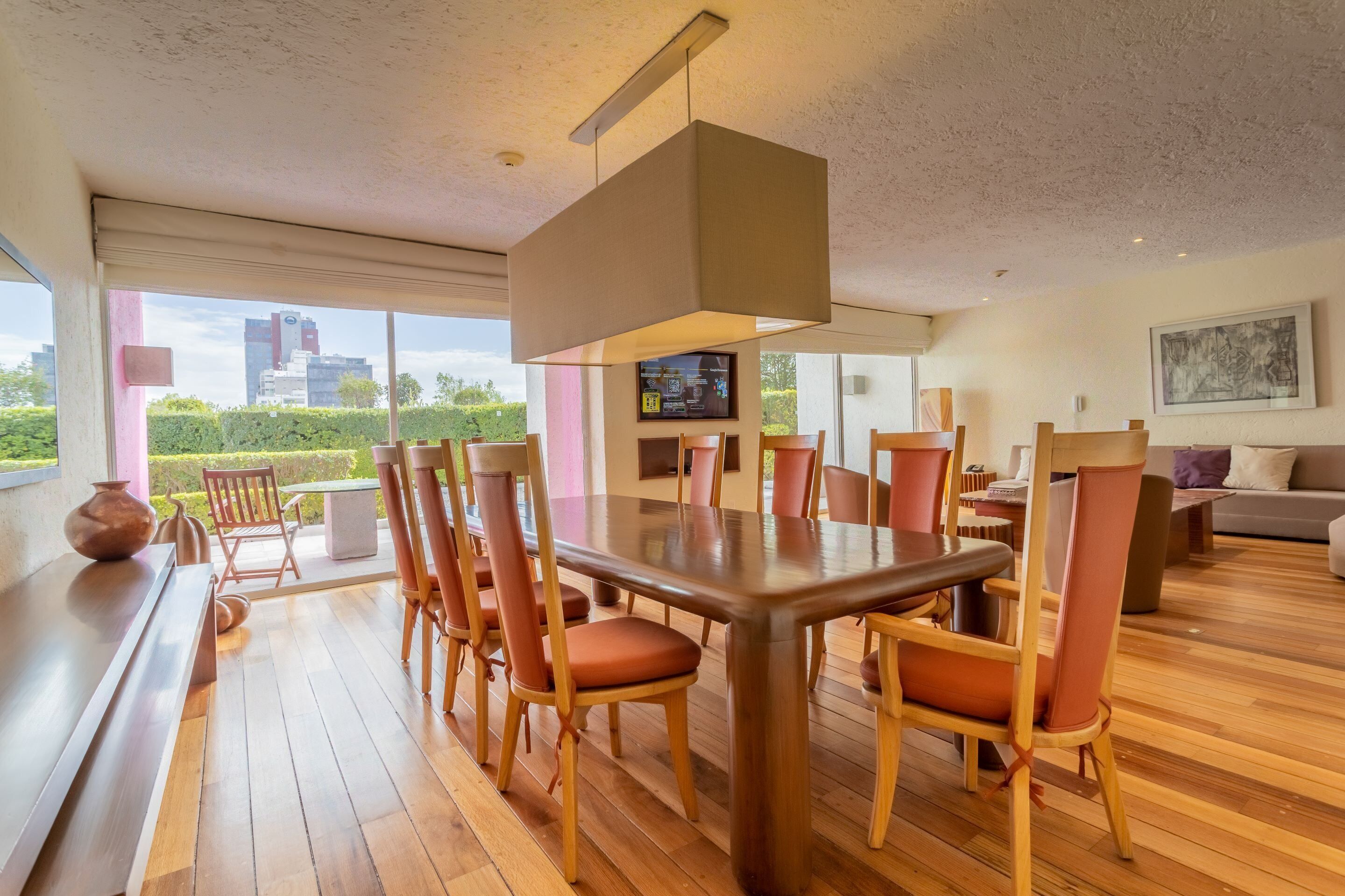Uma área de refeições moderna com uma grande mesa de madeira e cadeiras combinando, uma televisão na parede e vista para a cidade através de amplas janelas.