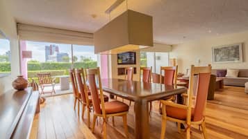 Uma área de refeições moderna com uma grande mesa de madeira e cadeiras combinando, uma televisão na parede e vista para a cidade através de amplas janelas.