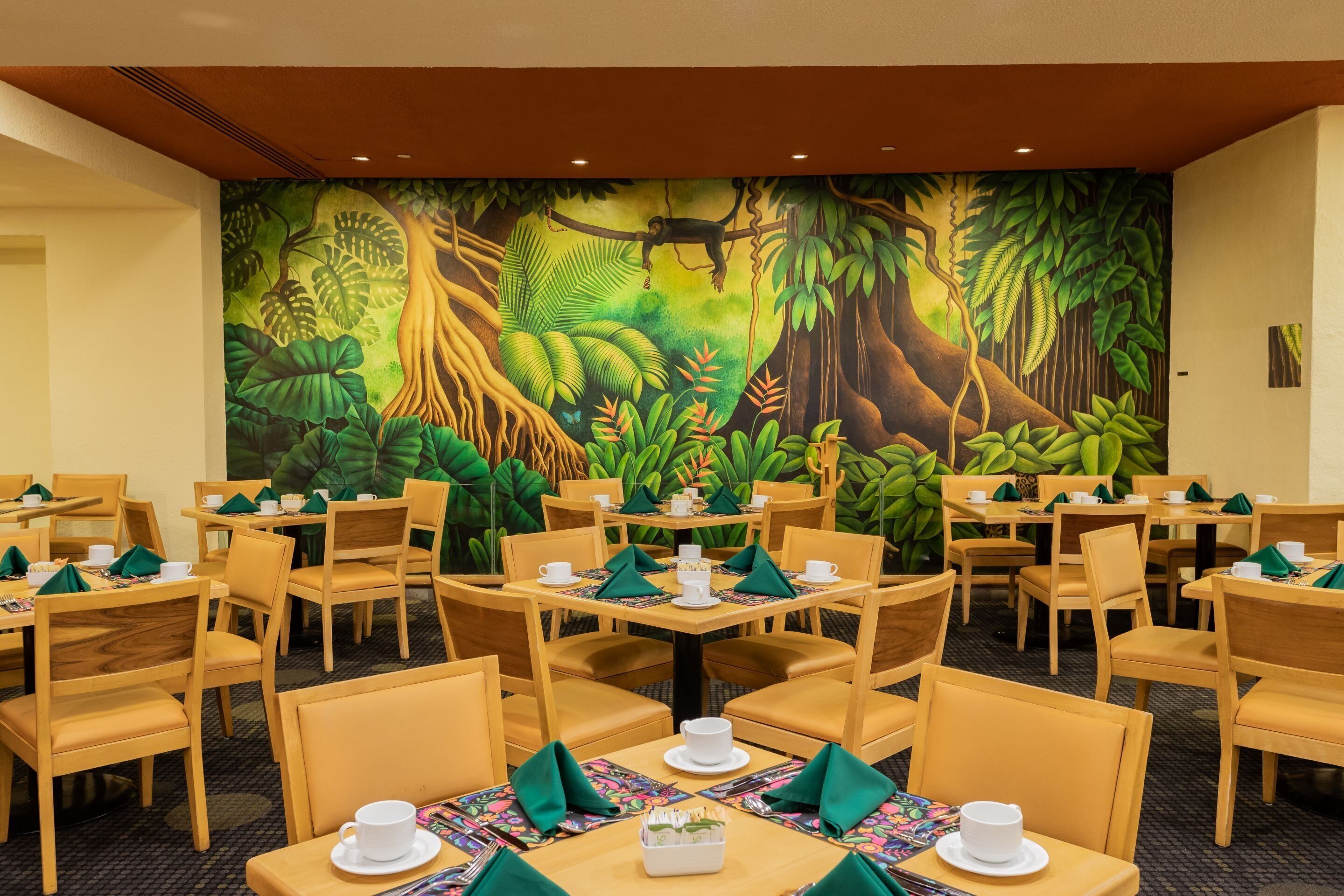 Un restaurante con mesas y sillas redondas, un mural de selva en la pared y servilletas verdes.