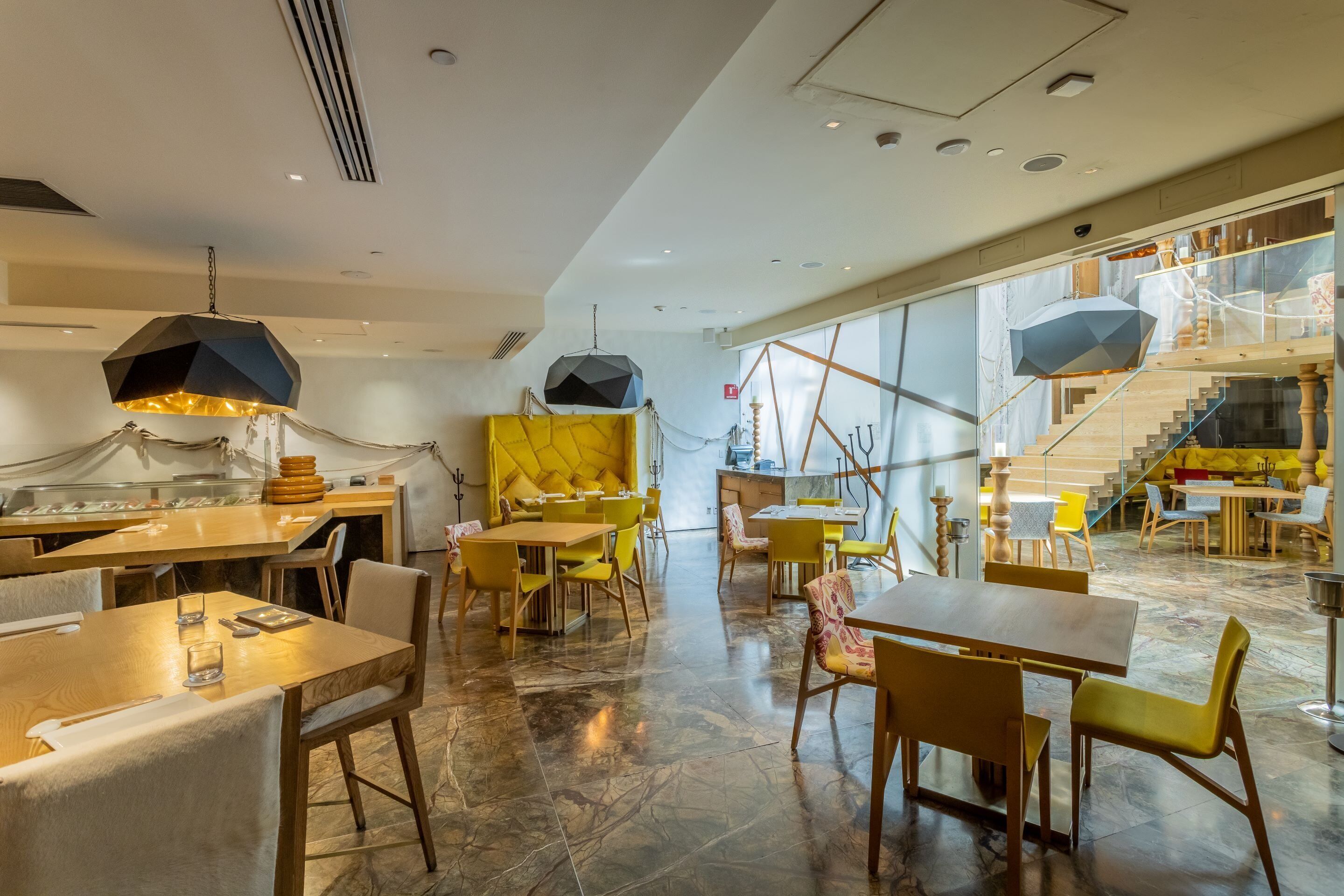 Ein modernes Restaurant mit Holztischen und gelben Stühlen, einem Küchenbereich und einer Treppe, die in ein Obergeschoss führt.