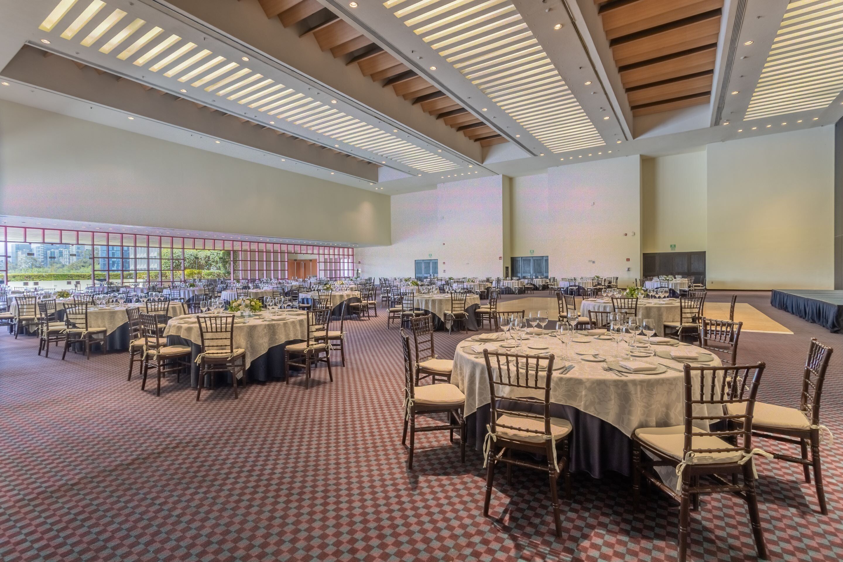 Une salle de banquet spacieuse, dotée de nombreuses tables rondes dressées pour le repas, avec un tapis à motifs et un design de plafond moderne.