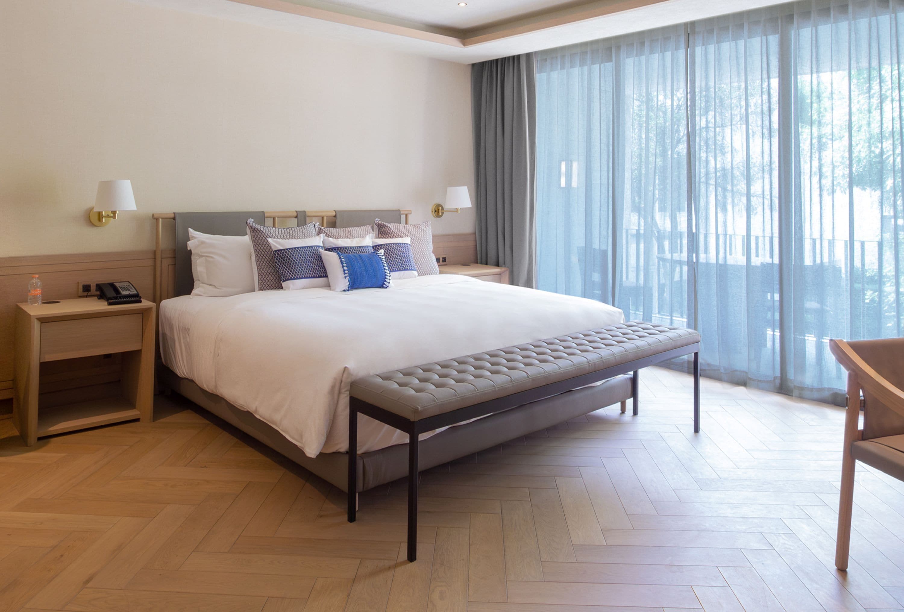 A hotel room with a large bed, two bedside tables, a bench, and a view of the outdoors through sheer curtains.