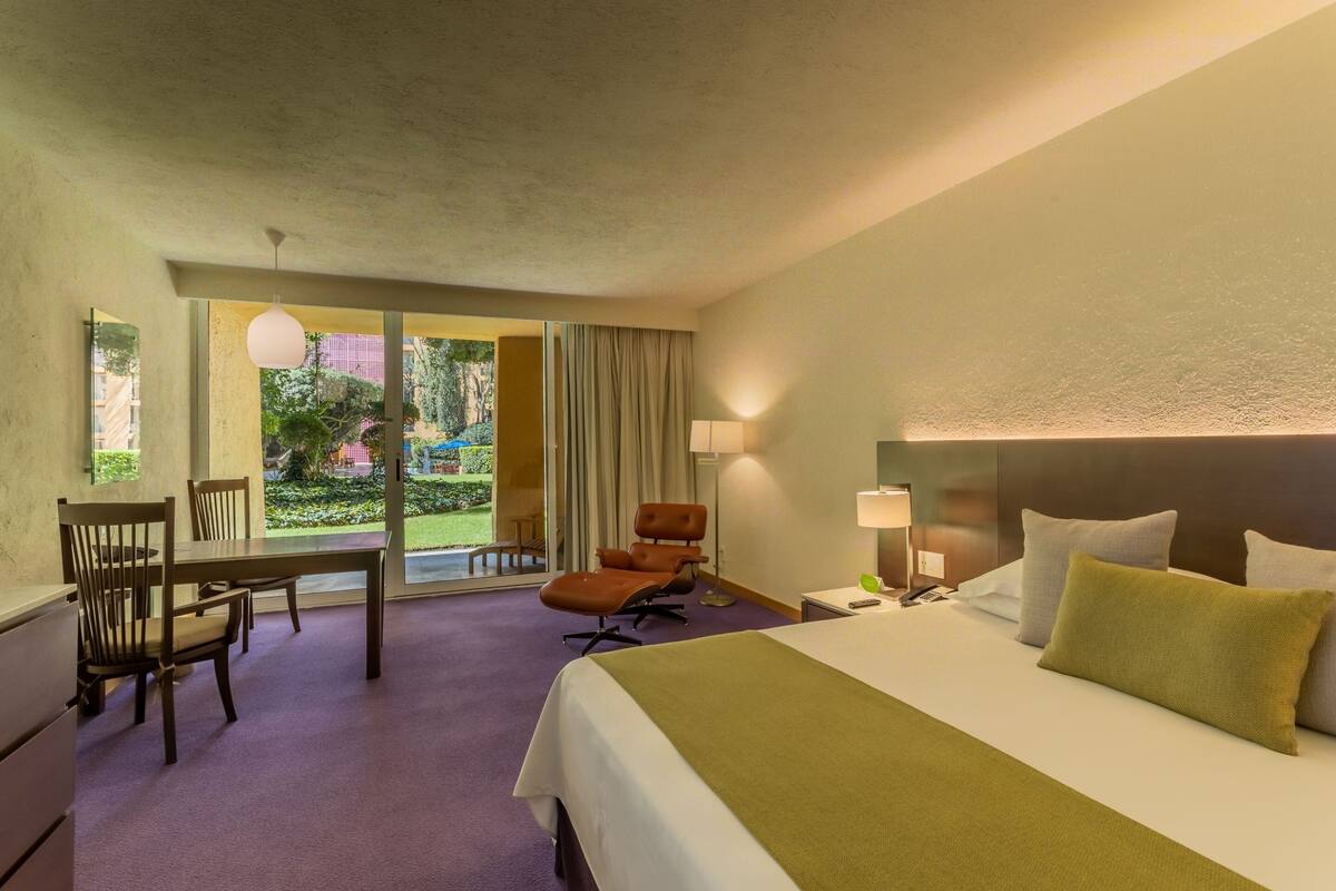 A hotel room with a bed, a desk, a chair, and a view of a garden through a glass door.